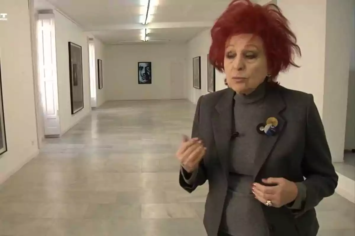 Una mujer con cabello rojo y un traje gris está de pie en una galería de arte, hablando mientras gesticula con la mano.