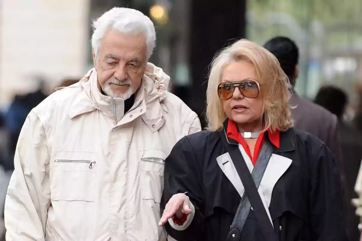 Una pareja de personas mayores caminando juntas al aire libre, el hombre lleva una chaqueta clara y la mujer usa gafas de sol grandes.