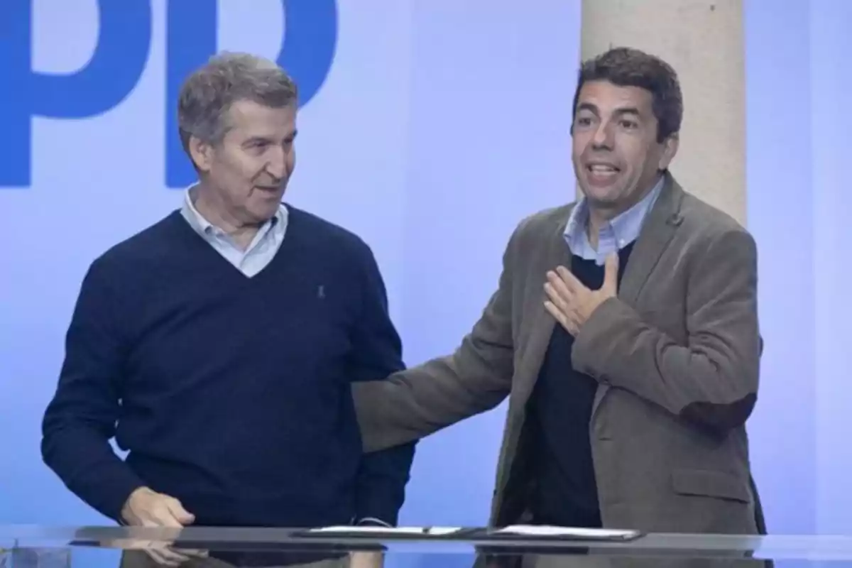 Dos hombres de pie en un escenario con fondo azul, uno de ellos con la mano en el pecho. Dos hombres de pie en un escenario con fondo azul, uno de ellos con la mano en el pecho.
