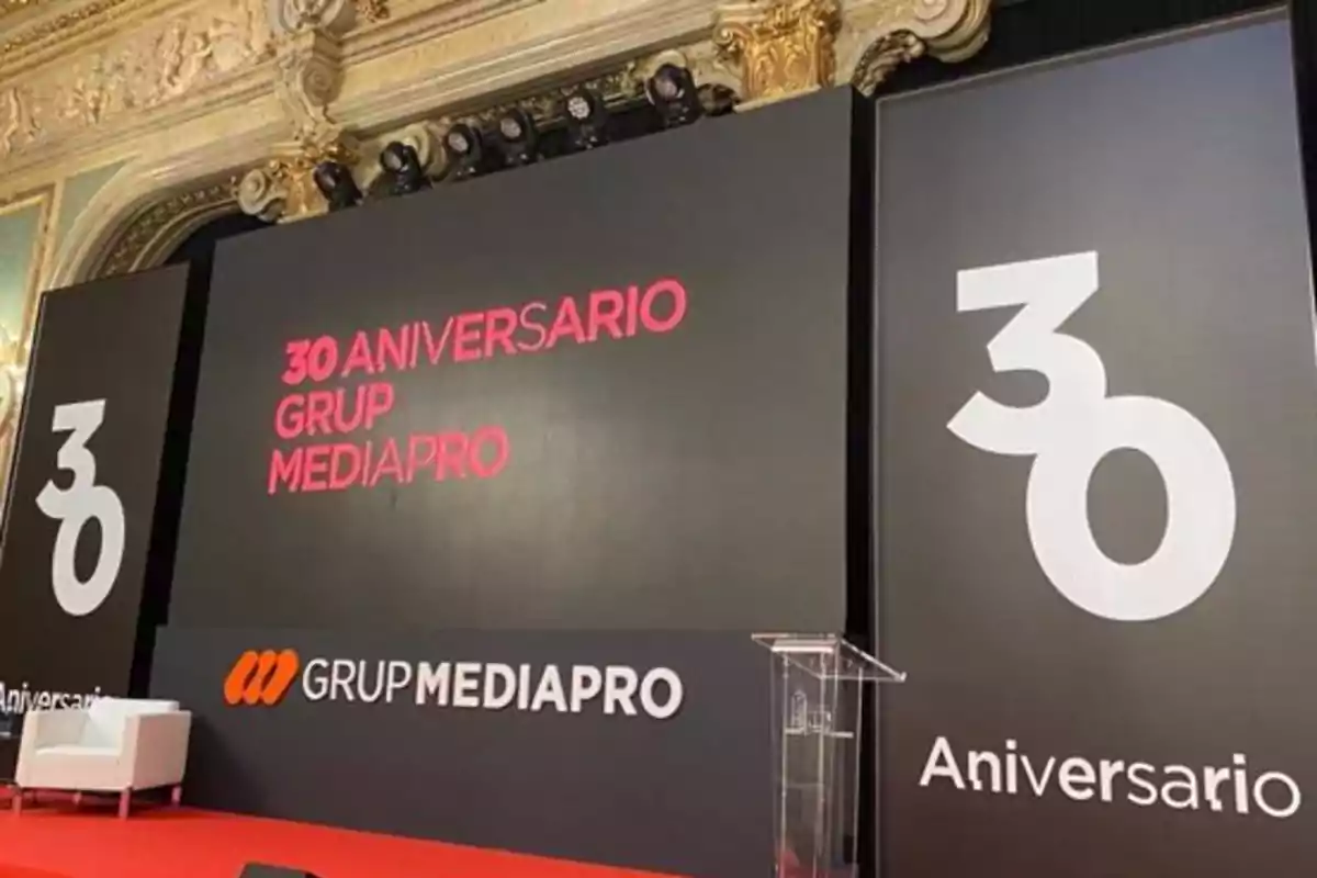 Escenario decorado para el 30 aniversario de Grup Mediapro con fondo negro, letras blancas y rojas, un sillón blanco y un atril transparente. Escenario decorado para el 30 aniversario de Grup Mediapro con fondo negro, letras blancas y rojas, un sillón blanco y un atril transparente.