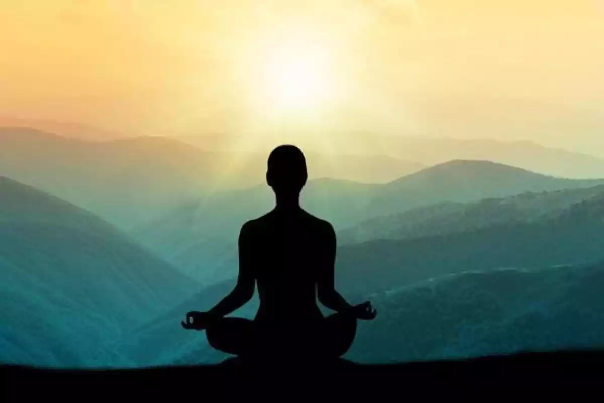 Persona meditando en posición de loto con un paisaje montañoso y un amanecer de fondo.
