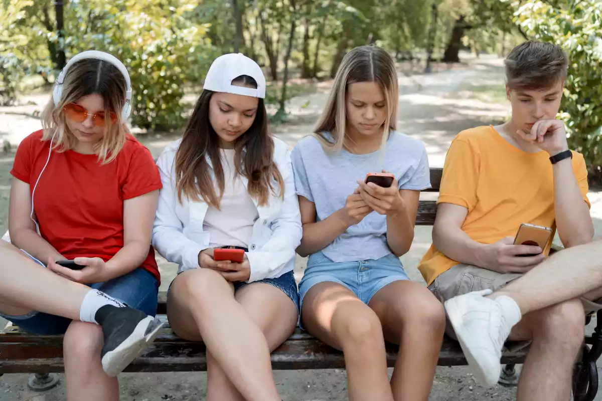 Cuatro jóvenes sentados en un banco al aire libre, cada uno concentrado en su teléfono móvil.