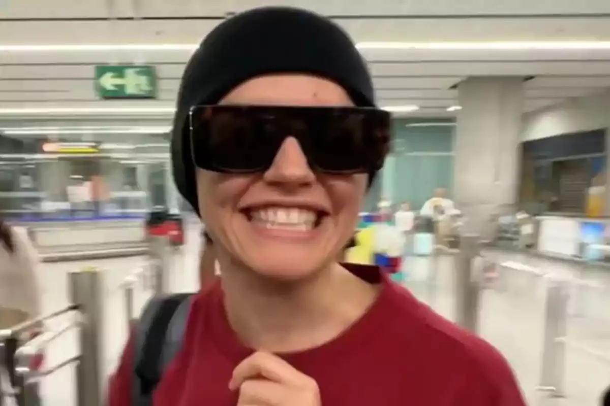 Una persona sonriente con gafas de sol grandes y un gorro negro en un entorno de aeropuerto. Una persona sonriente con gafas de sol grandes y un gorro negro en un entorno de aeropuerto.