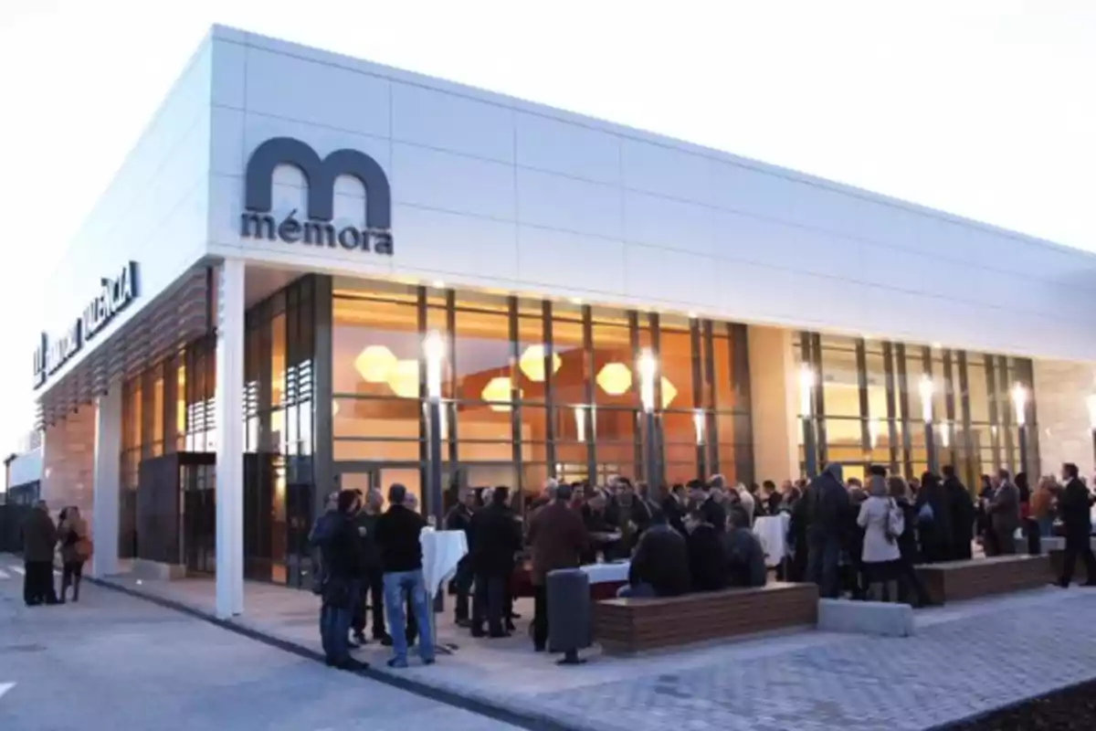 Un grupo de personas se reúne frente a un edificio moderno con grandes ventanales y un letrero que dice Un grupo de personas se reúne frente a un edificio moderno con grandes ventanales y un letrero que dice