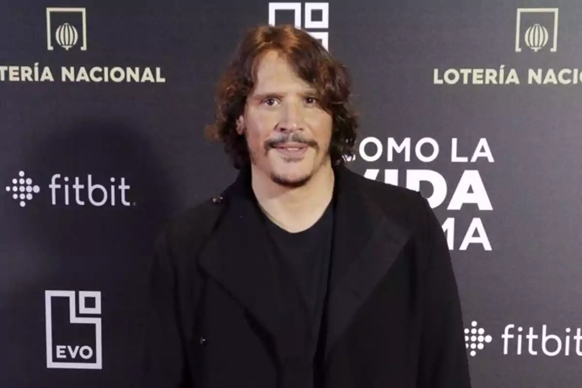 Hombre de cabello ondulado y bigote posando en un evento con fondo de logotipos y letras blancas sobre fondo negro Hombre de cabello ondulado y bigote posando en un evento con fondo de logotipos y letras blancas sobre fondo negro