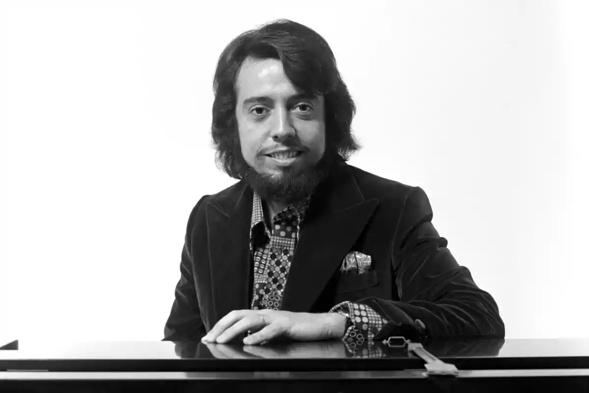 Un hombre con barba y cabello largo, vestido con un traje de terciopelo, sentado frente a un piano.