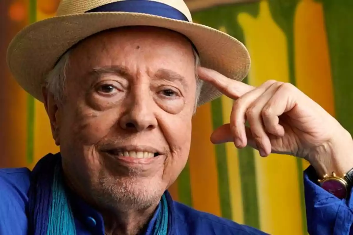 Hombre mayor con sombrero de paja y camisa azul sonriendo frente a un fondo colorido.