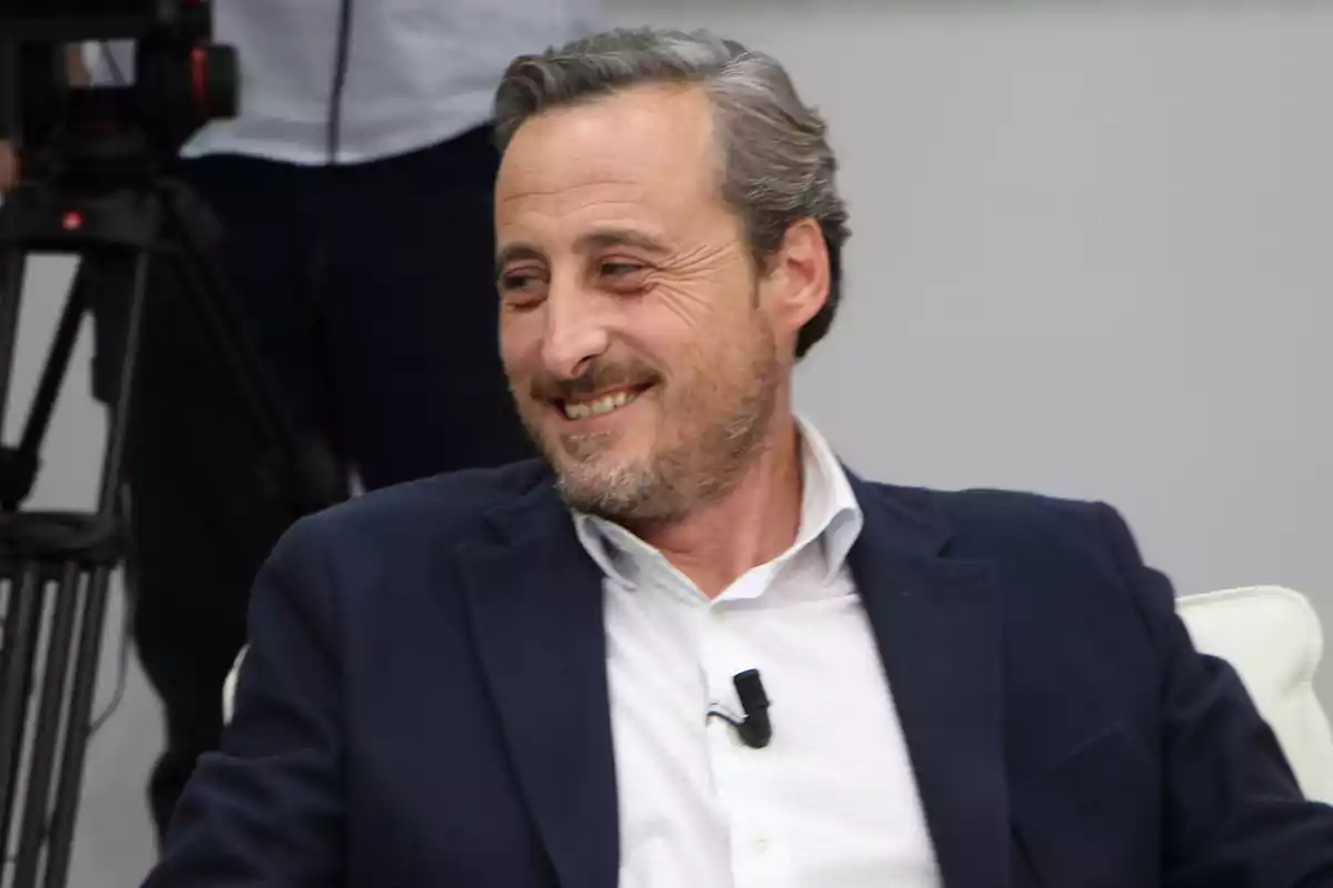 Un hombre de mediana edad con barba y cabello canoso sonríe mientras está sentado, lleva un traje oscuro y una camisa clara. Un hombre de mediana edad con barba y cabello canoso sonríe mientras está sentado, lleva un traje oscuro y una camisa clara.