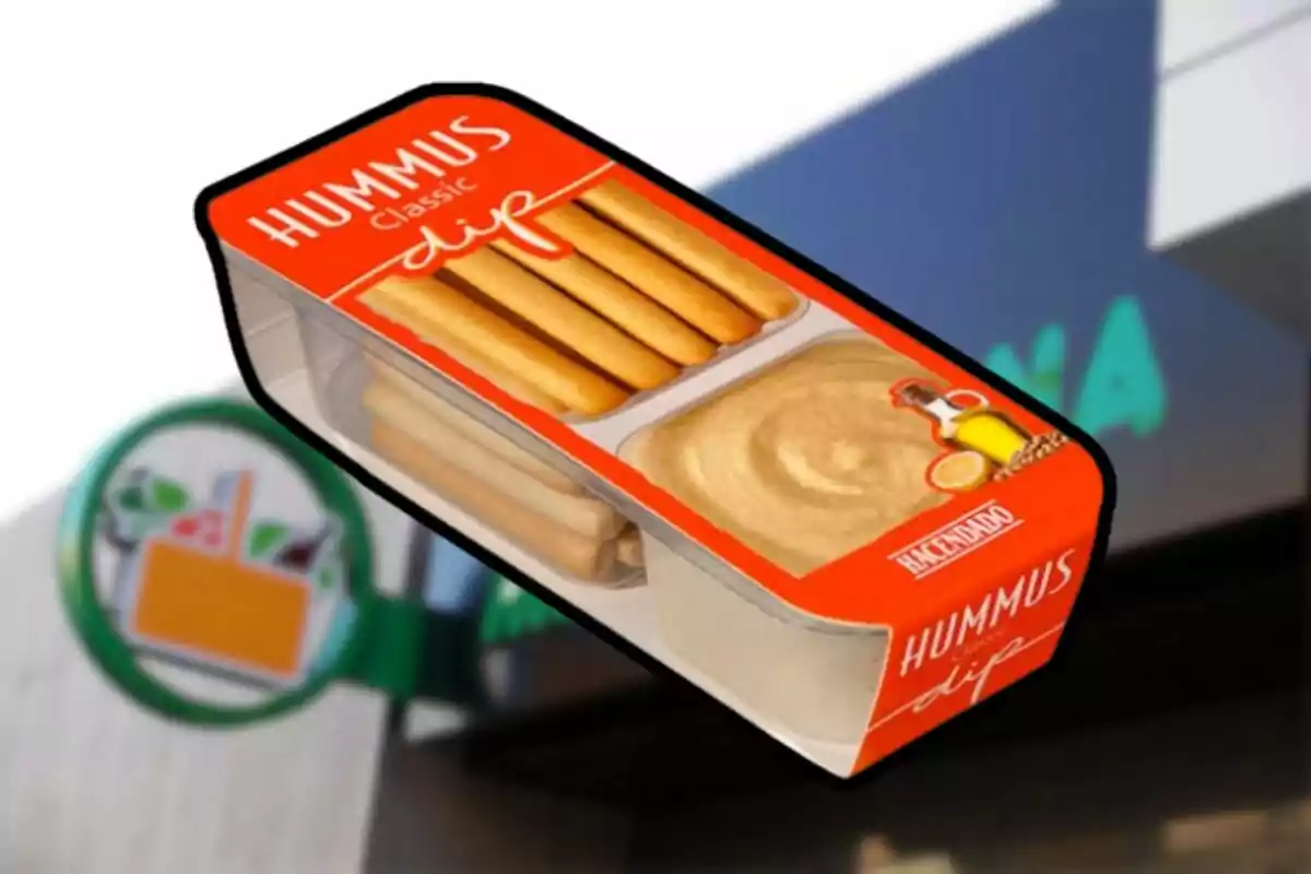 Envase de hummus clásico con palitos de pan de la marca Hacendado frente a un cartel de supermercado. Envase de hummus clásico con palitos de pan de la marca Hacendado frente a un cartel de supermercado.