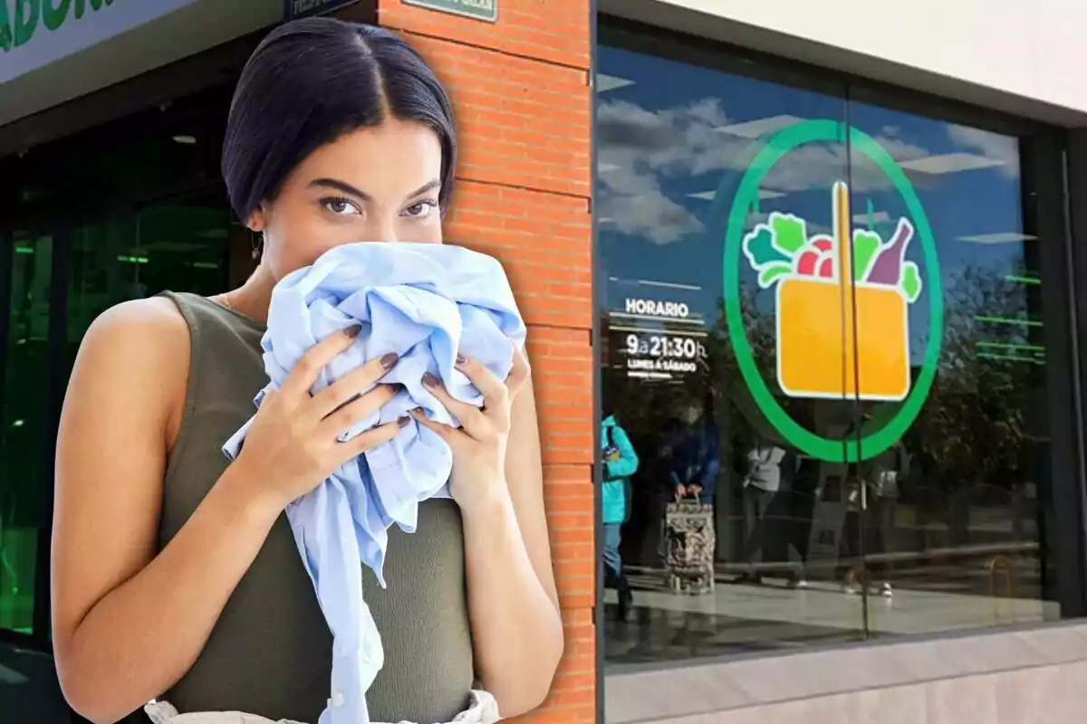 Mujer sosteniendo y oliendo una prenda azul frente a la entrada de un supermercado con un logo de cesta de compras en la ventana