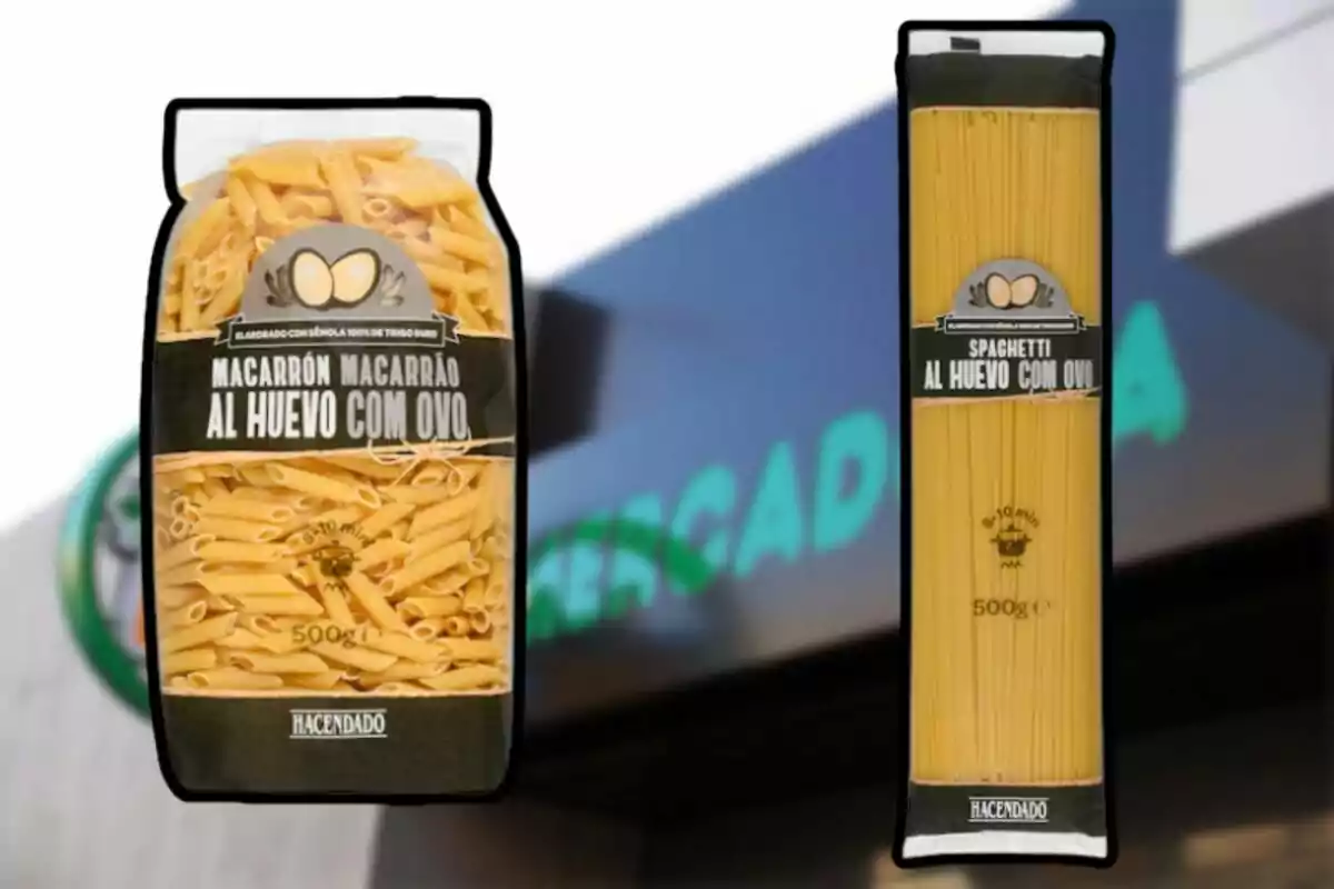 Dos paquetes de pasta Hacendado, uno de macarrones y otro de espaguetis al huevo, frente a un cartel borroso de supermercado. Dos paquetes de pasta Hacendado, uno de macarrones y otro de espaguetis al huevo, frente a un cartel borroso de supermercado.