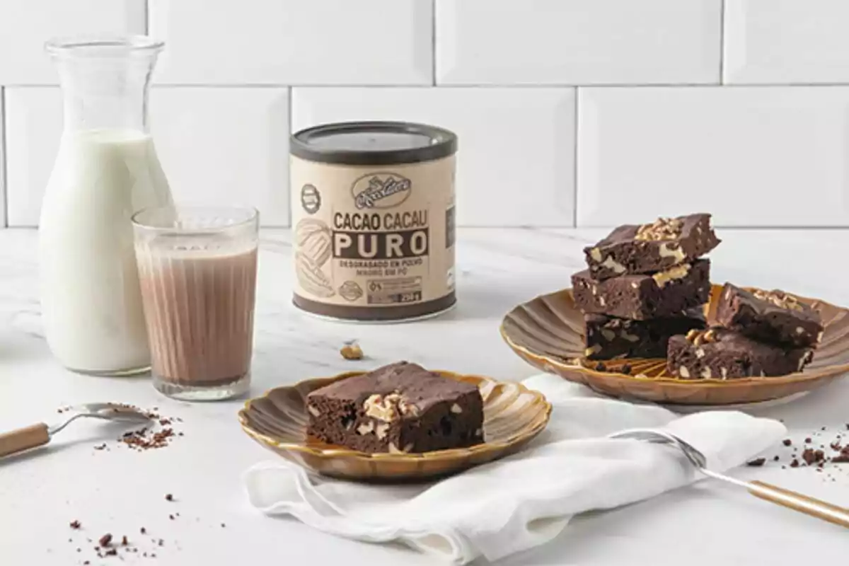 Una jarra de leche junto a un vaso de leche con cacao en polvo y un plato con brownies de chocolate sobre una mesa blanca. Una jarra de leche junto a un vaso de leche con cacao en polvo y un plato con brownies de chocolate sobre una mesa blanca.