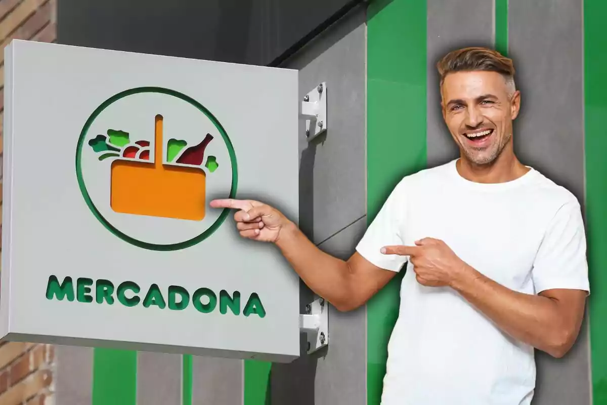 Un hombre sonriente con camiseta blanca señala el letrero de Mercadona en una pared.