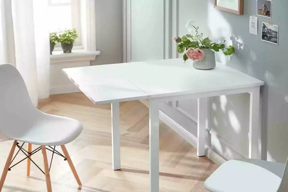 Una mesa blanca con una silla blanca en una habitación luminosa con piso de madera, una ventana con cortinas blancas y plantas en el alféizar, y una pared gris con decoraciones y un jarrón con flores sobre la mesa. Una mesa blanca con una silla blanca en una habitación luminosa con piso de madera, una ventana con cortinas blancas y plantas en el alféizar, y una pared gris con decoraciones y un jarrón con flores sobre la mesa.