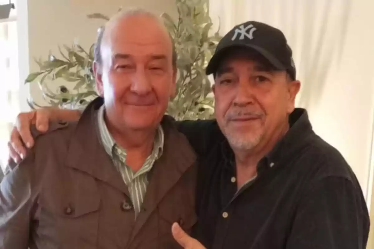 Dos hombres posan juntos sonriendo mientras uno de ellos lleva una gorra de béisbol.