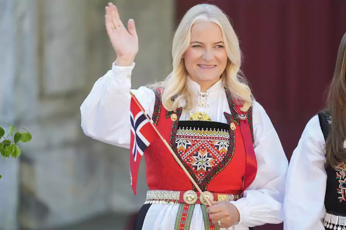 Mette-Marit con un traje tradicional noruego sostiene una bandera mientras sonríe y saluda. Mette-Marit con un traje tradicional noruego sostiene una bandera mientras sonríe y saluda.
