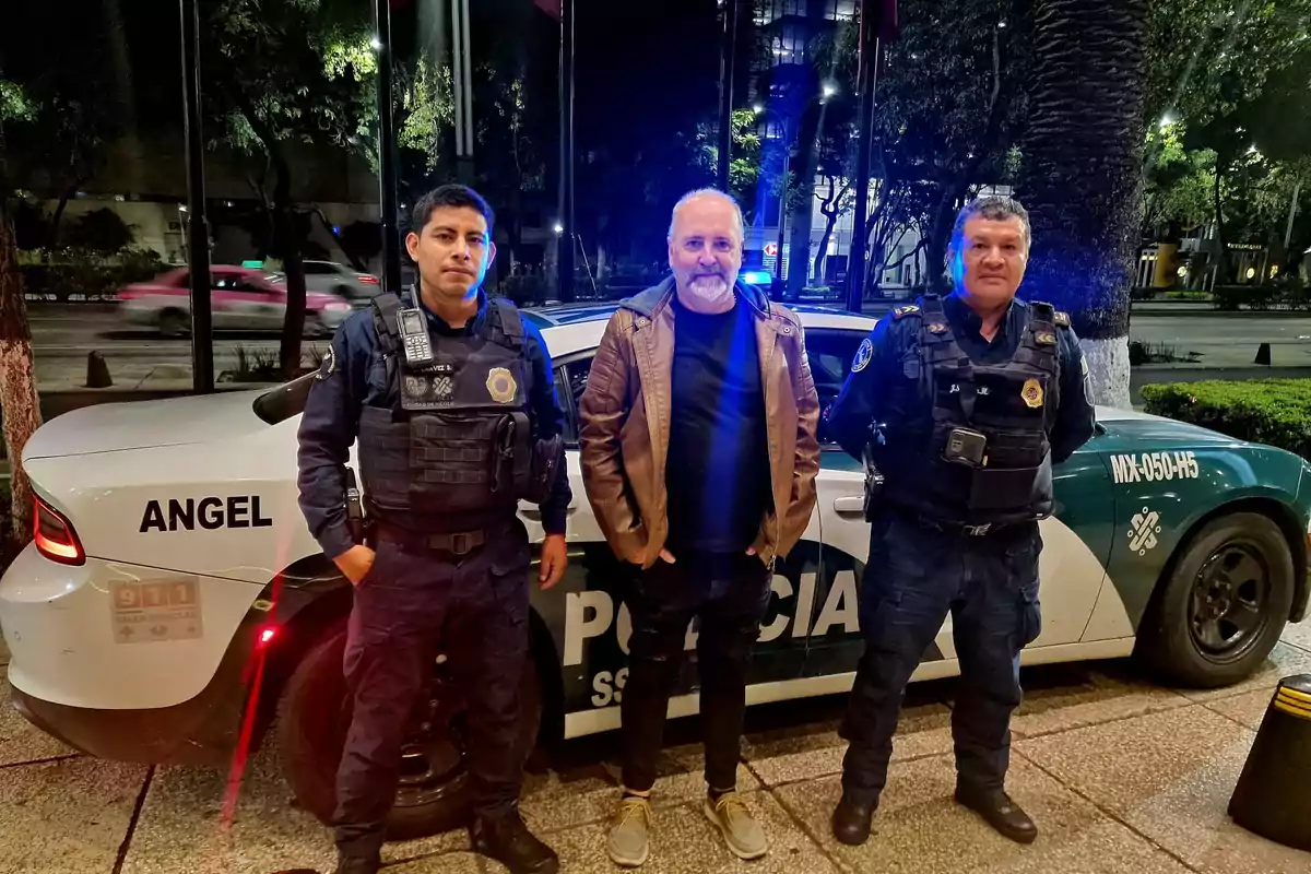 Un hombre de pie entre dos policías frente a un coche patrulla en una calle iluminada por la noche.