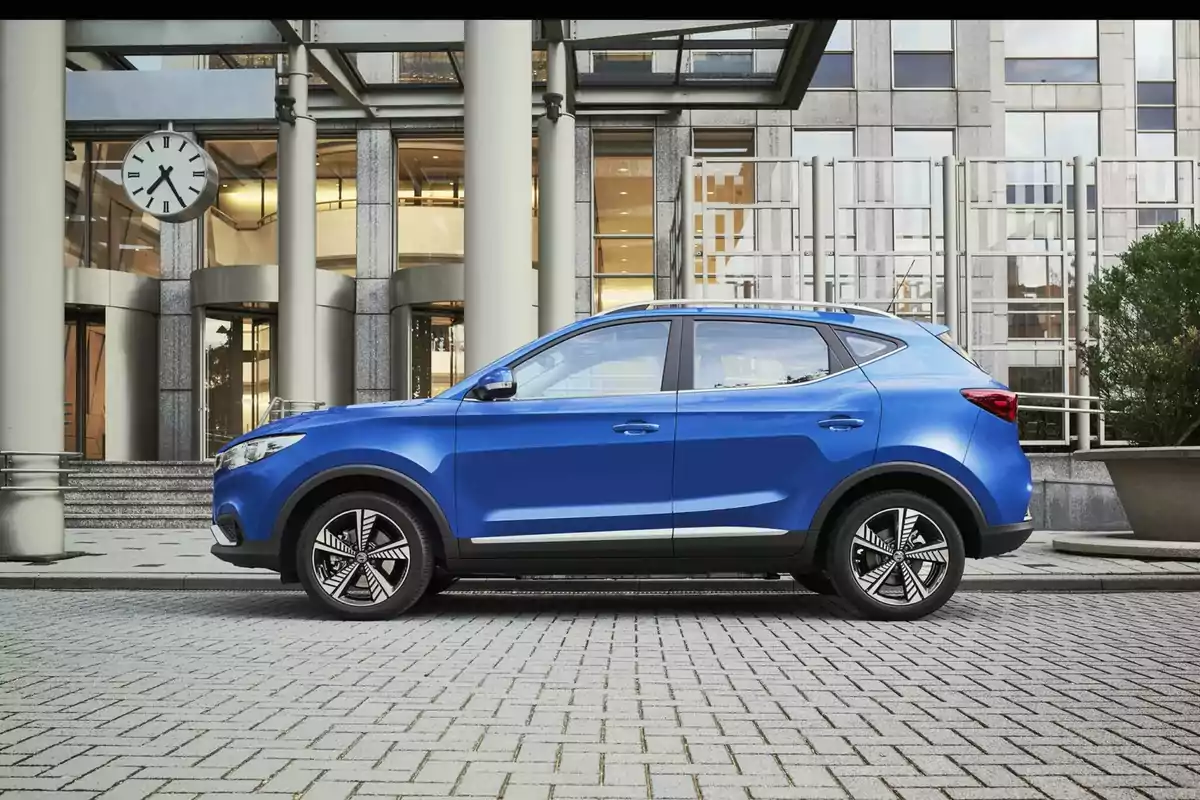 Un MG ZS EV azul estacionado frente a un edificio moderno con un reloj grande en el fondo.