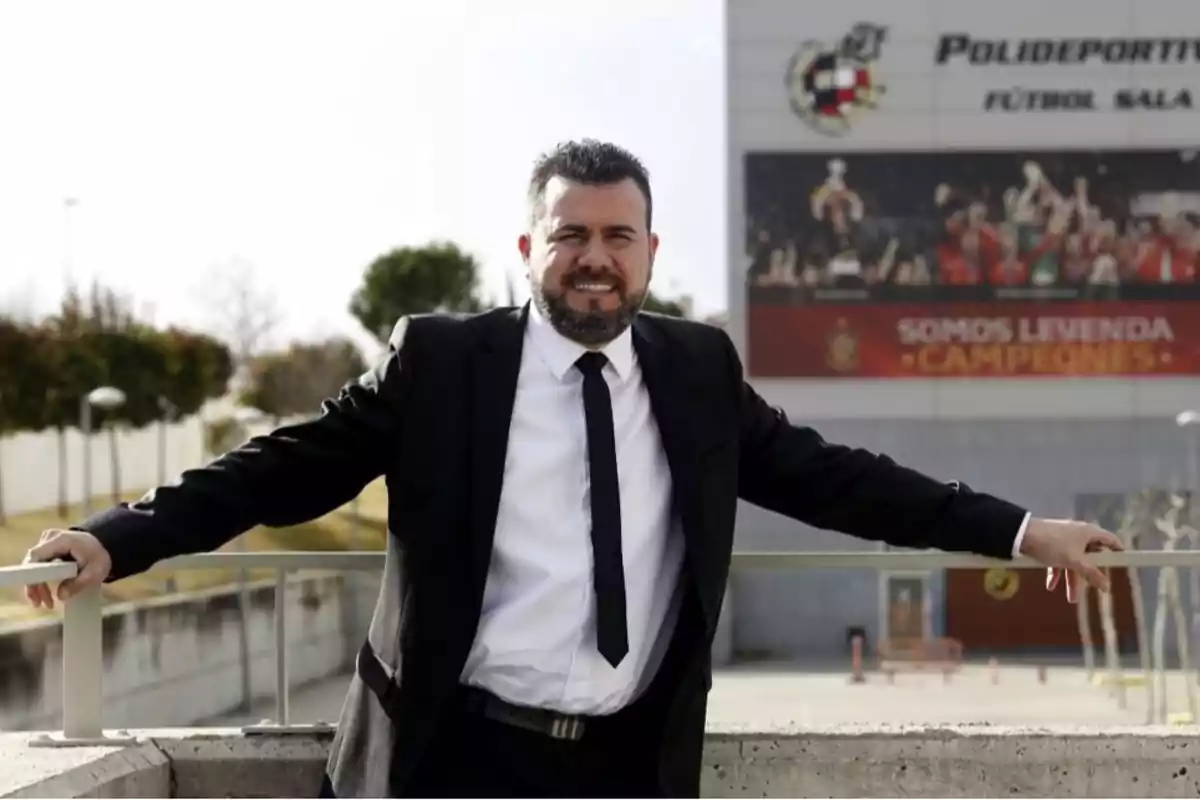 Un hombre con traje y corbata sonríe mientras se apoya en una barandilla al aire libre con un edificio deportivo de fondo. Un hombre con traje y corbata sonríe mientras se apoya en una barandilla al aire libre con un edificio deportivo de fondo.