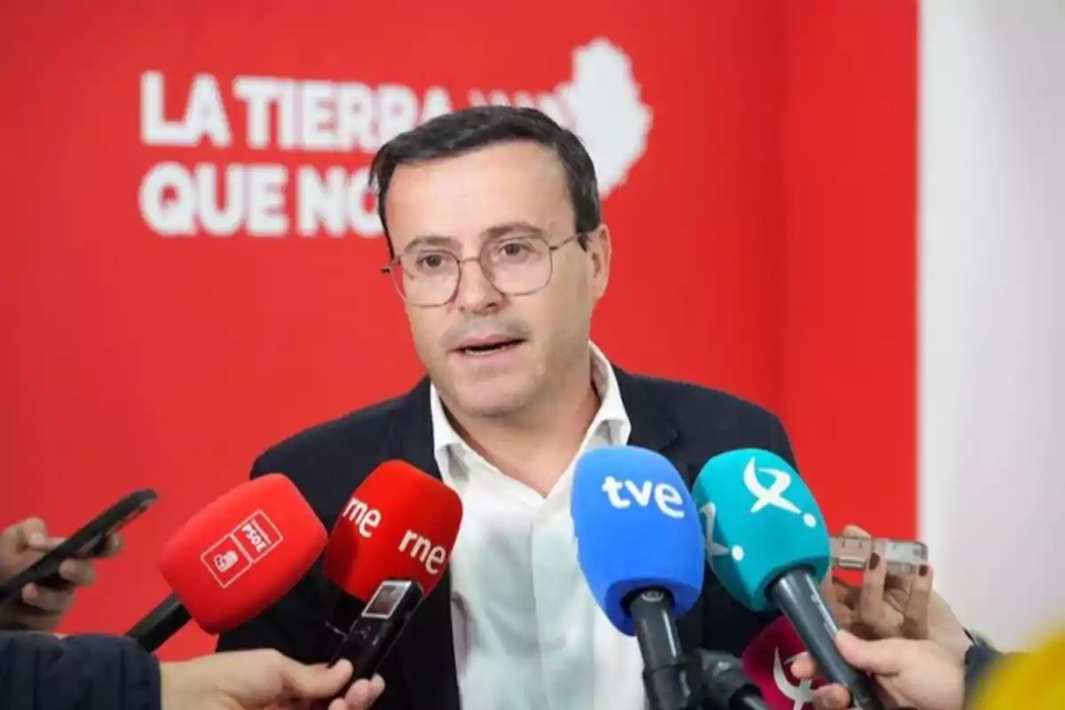 Hombre con gafas y chaqueta oscura hablando ante varios micrófonos de diferentes medios de comunicación con fondo rojo y letras blancas Hombre con gafas y chaqueta oscura hablando ante varios micrófonos de diferentes medios de comunicación con fondo rojo y letras blancas