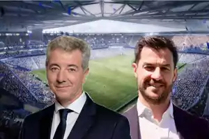 Dos hombres posan frente a un estadio de fútbol lleno de espectadores.