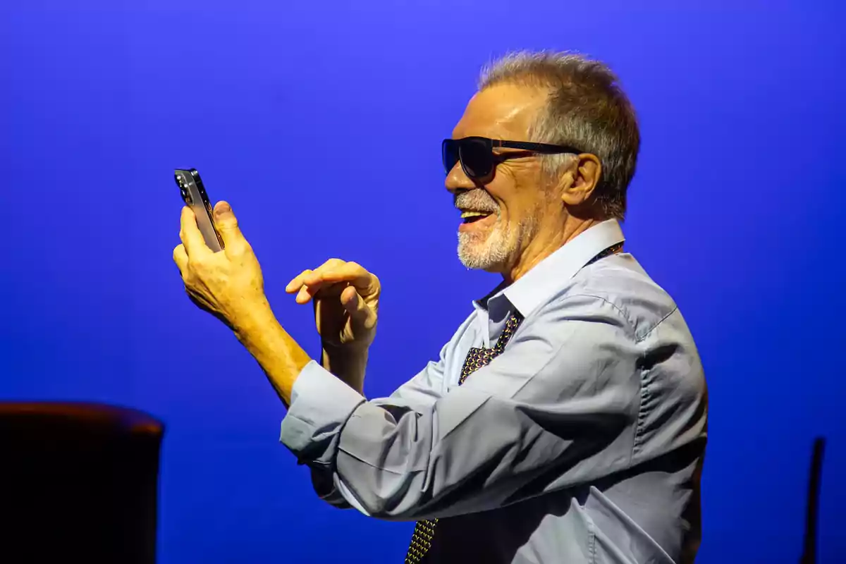 Hombre mayor con gafas de sol sonriendo mientras usa un teléfono móvil frente a un fondo azul.