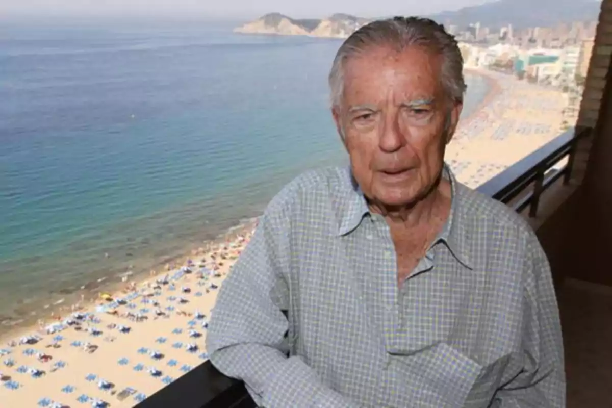 Un hombre mayor en un balcón con vista a una playa concurrida y el mar al fondo.