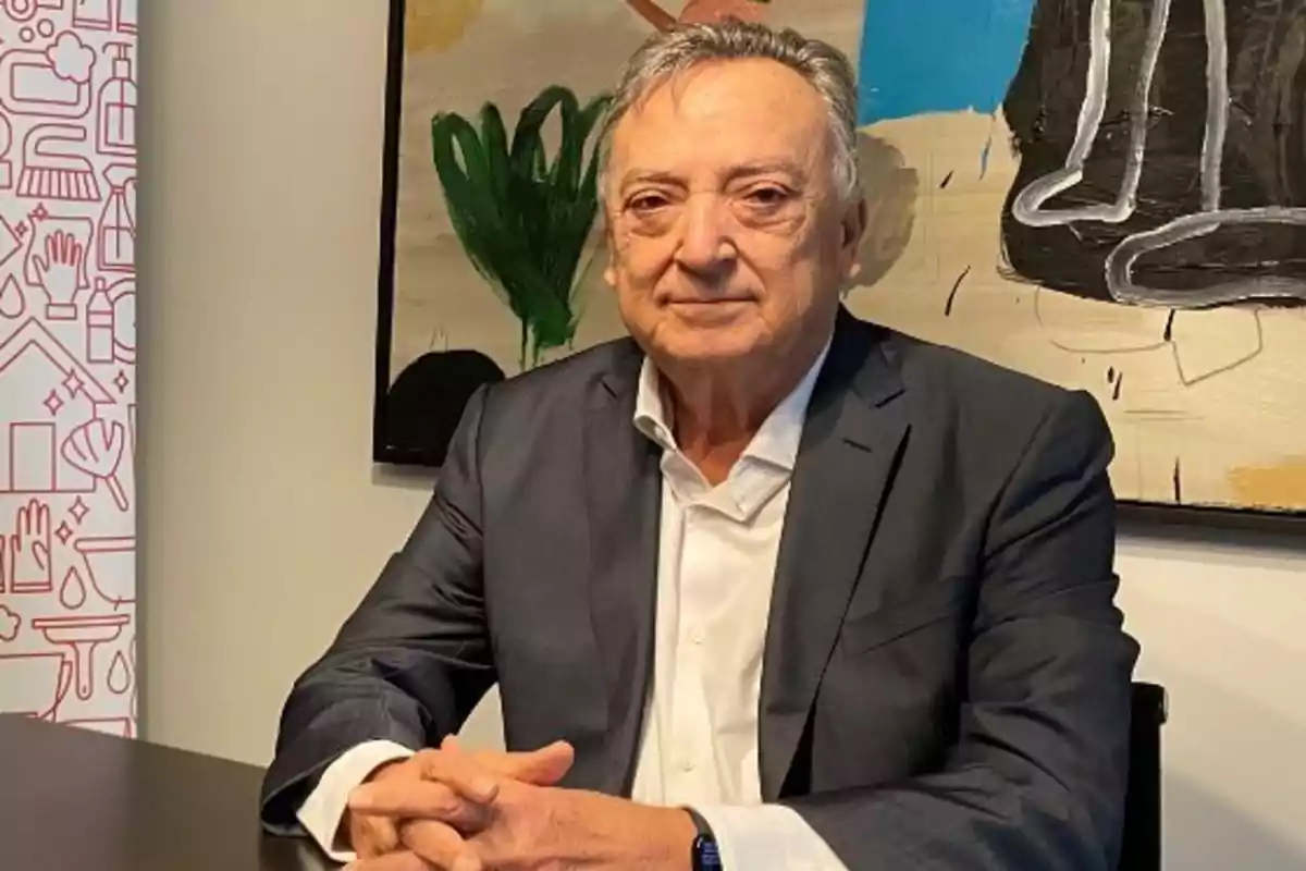 Un hombre mayor con traje oscuro y camisa blanca está sentado frente a una mesa con un cuadro colorido de fondo.