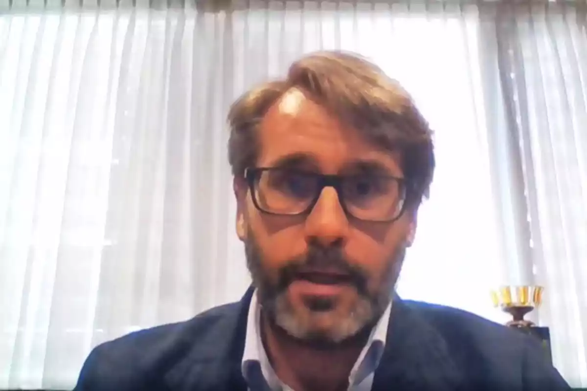 Un hombre con gafas y barba aparece en una videollamada con cortinas blancas de fondo. Un hombre con gafas y barba aparece en una videollamada con cortinas blancas de fondo.