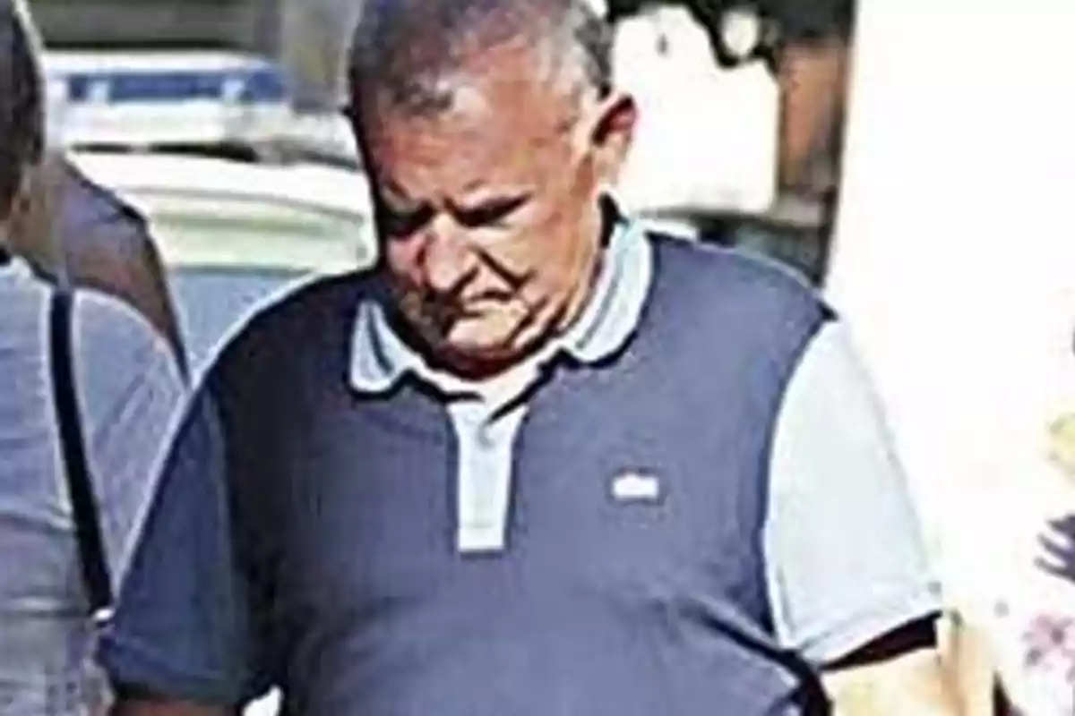 Un hombre mayor caminando con la cabeza baja y vistiendo una camiseta polo azul.