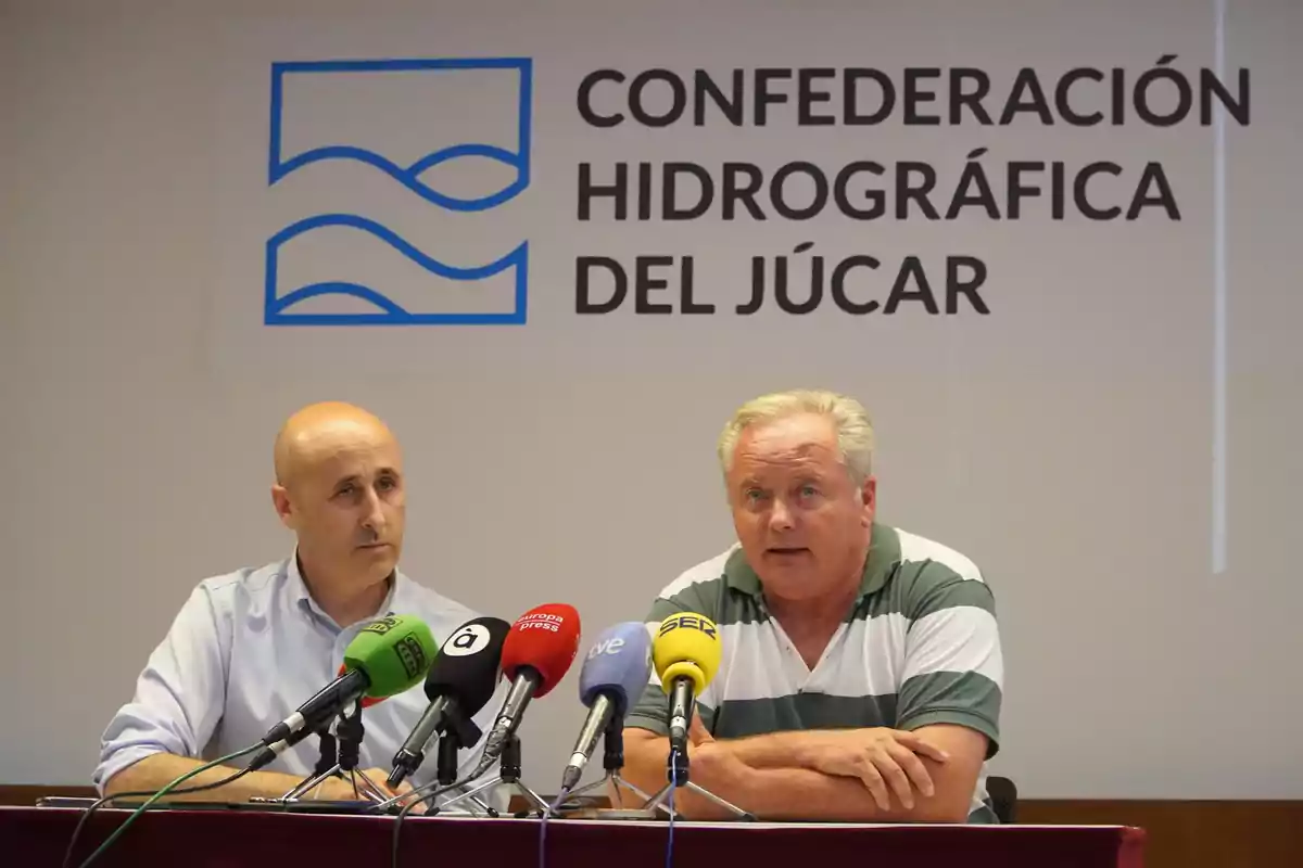 Dos hombres en una conferencia de prensa frente a varios micrófonos con el logotipo y nombre de la Confederación Hidrográfica del Júcar en el fondo.