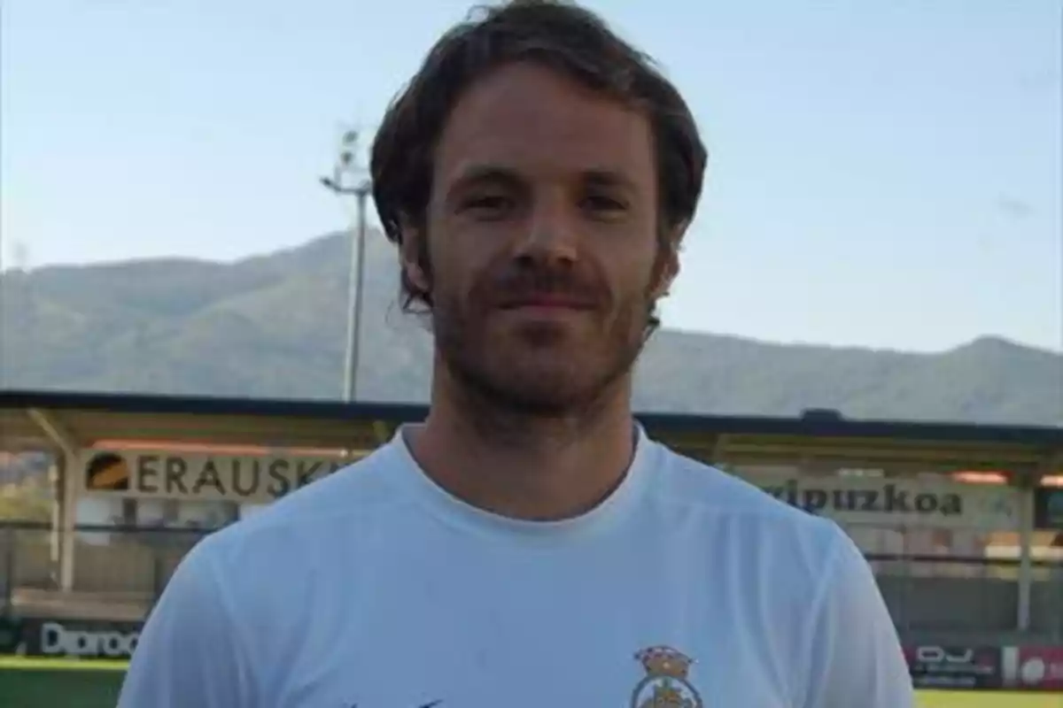 Hombre con camiseta blanca de fútbol posando en un campo con montañas al fondo Hombre con camiseta blanca de fútbol posando en un campo con montañas al fondo