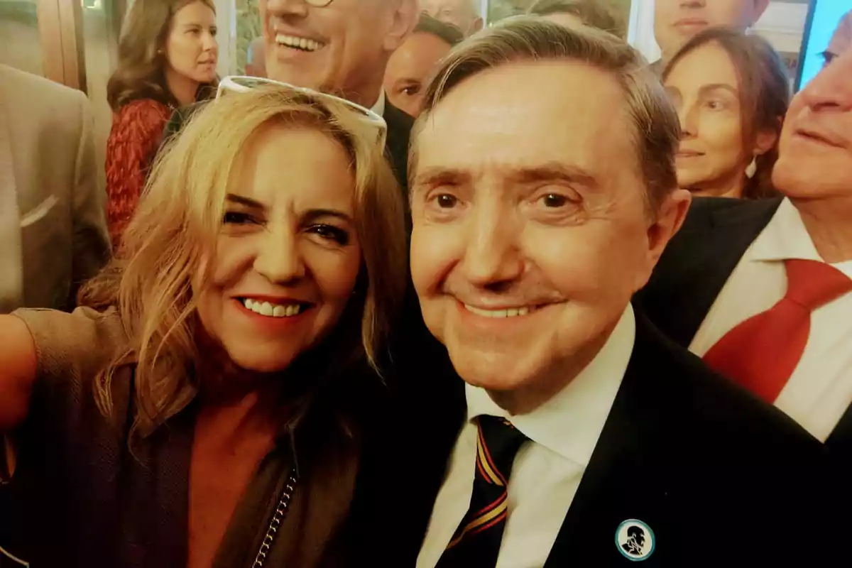 Una mujer y un hombre sonríen mientras se toman una selfie en un evento social rodeados de otras personas elegantemente vestidas Una mujer y un hombre sonríen mientras se toman una selfie en un evento social rodeados de otras personas elegantemente vestidas