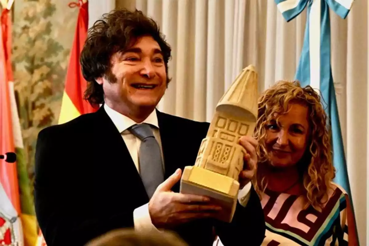 Hombre sonriente con traje sosteniendo un trofeo de madera en forma de edificio junto a una mujer rubia con cabello rizado