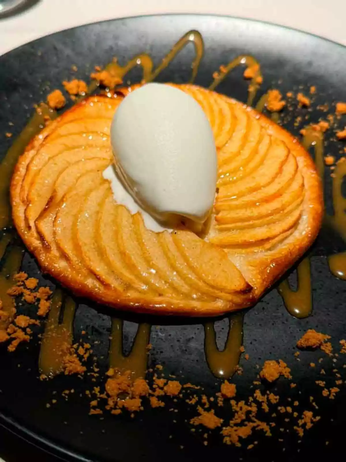 Un postre de tarta de manzana con una bola de helado de vainilla sobre un plato negro, decorado con caramelo y migas de galleta.