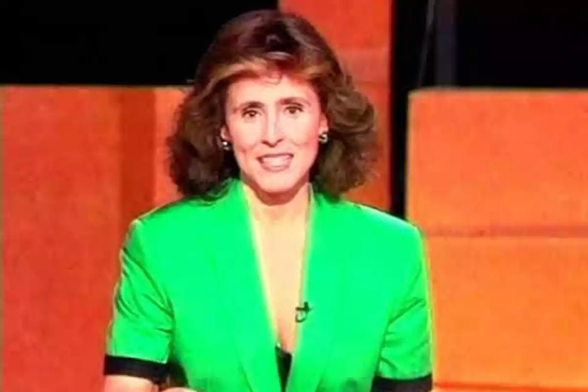 Una persona con cabello oscuro y rizado lleva un traje verde brillante y está sonriendo frente a un fondo naranja. Una persona con cabello oscuro y rizado lleva un traje verde brillante y está sonriendo frente a un fondo naranja.