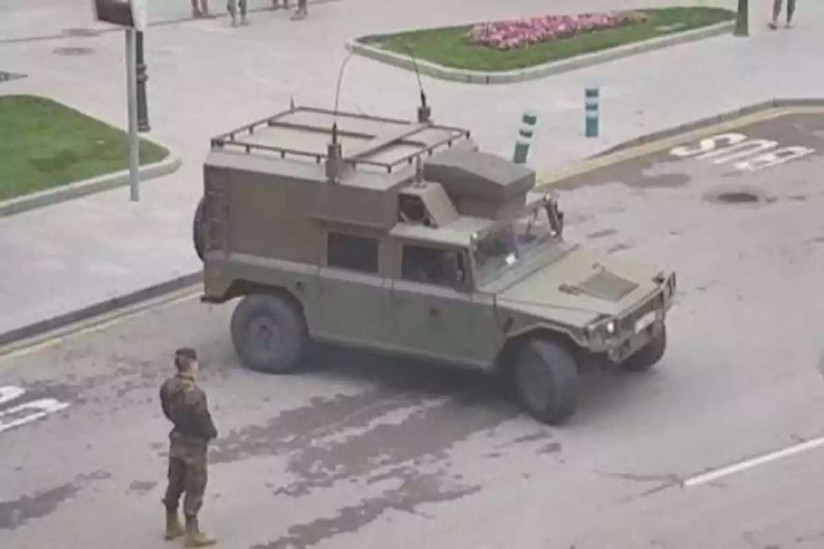 Un vehículo militar verde está estacionado en una calle mientras un soldado de pie observa.