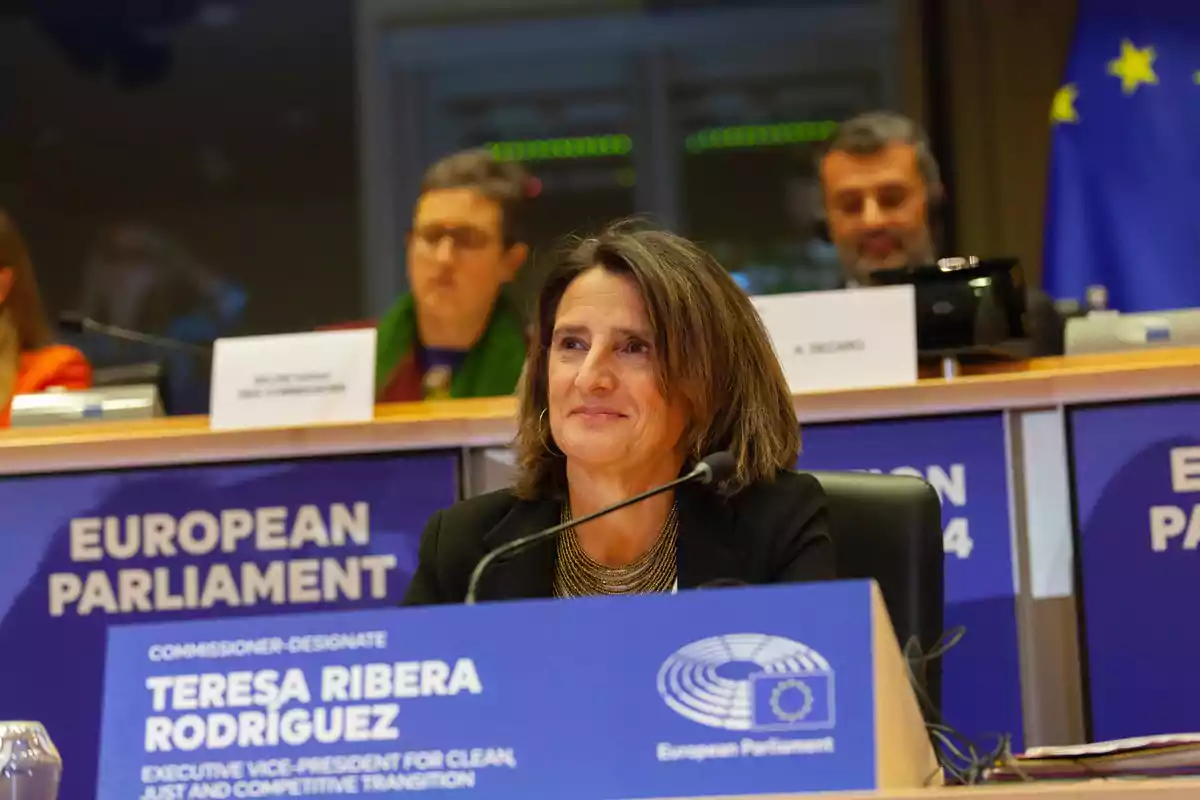 Una mujer sentada en una mesa con un cartel que dice "European Parliament" y "Teresa Ribera Rodríguez" en un entorno de conferencia.