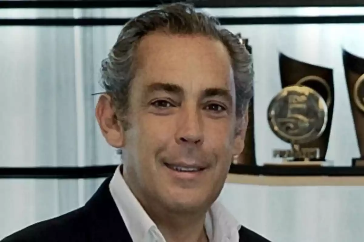 Hombre de cabello canoso y corto vistiendo saco oscuro y camisa blanca con trofeos en el fondo