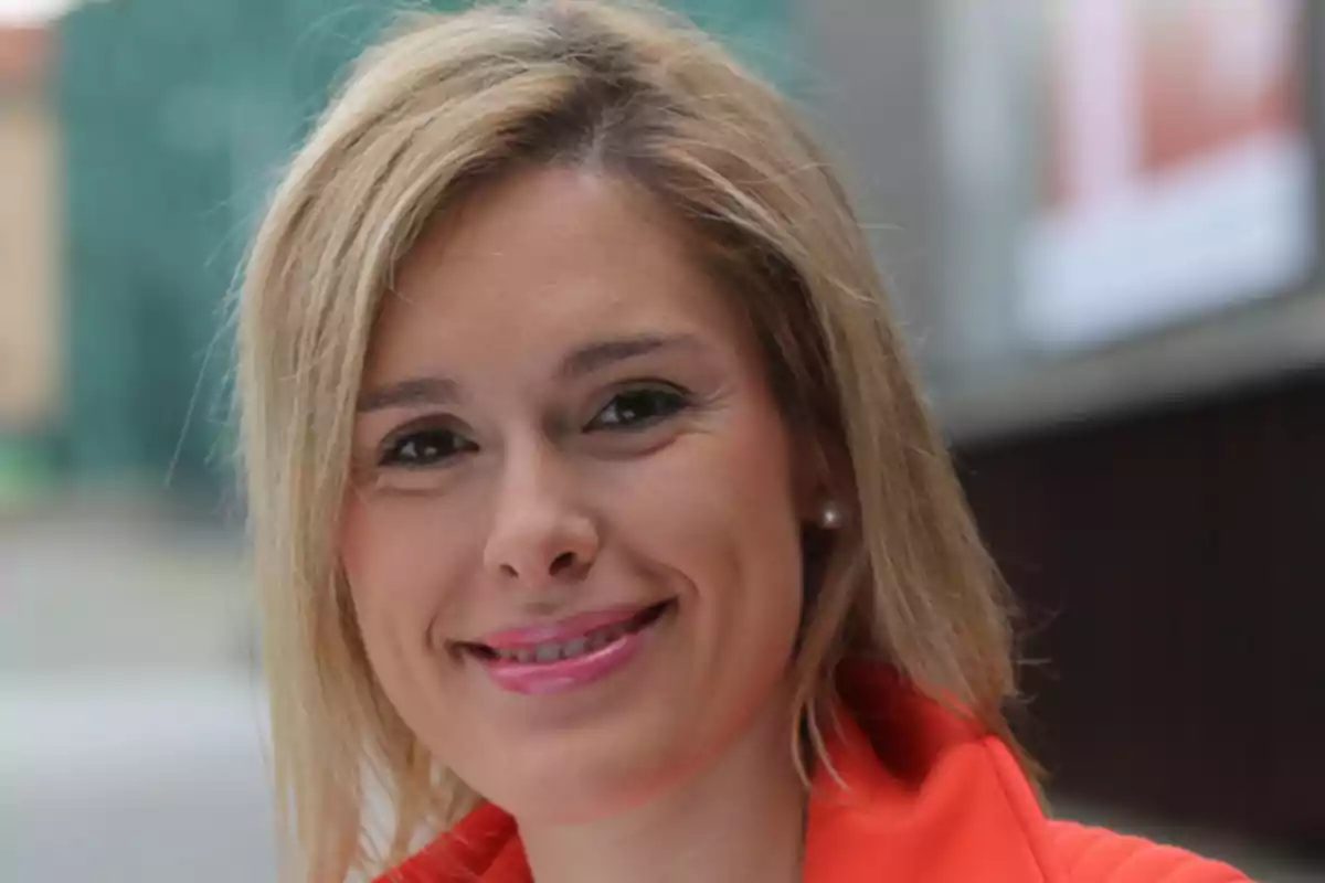 Una persona sonriendo con cabello rubio y chaqueta roja.