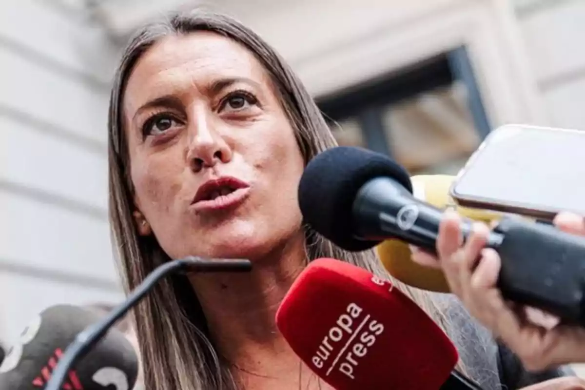 Mujer hablando frente a varios micrófonos de prensa en un entorno exterior Mujer hablando frente a varios micrófonos de prensa en un entorno exterior