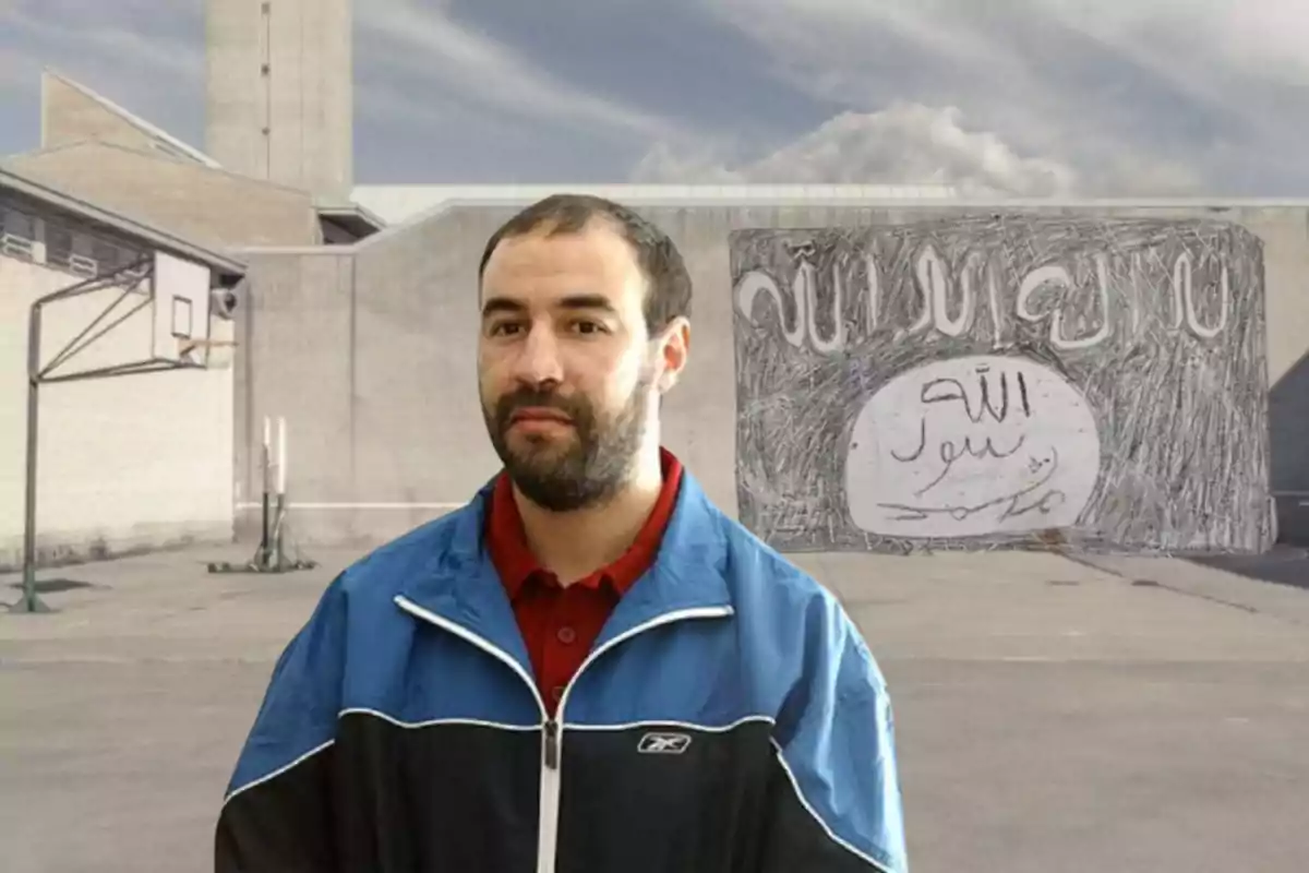 Un hombre con chaqueta azul y roja está de pie frente a una cancha de baloncesto al aire libre, con un edificio de fondo y un cartel con escritura árabe en la pared.