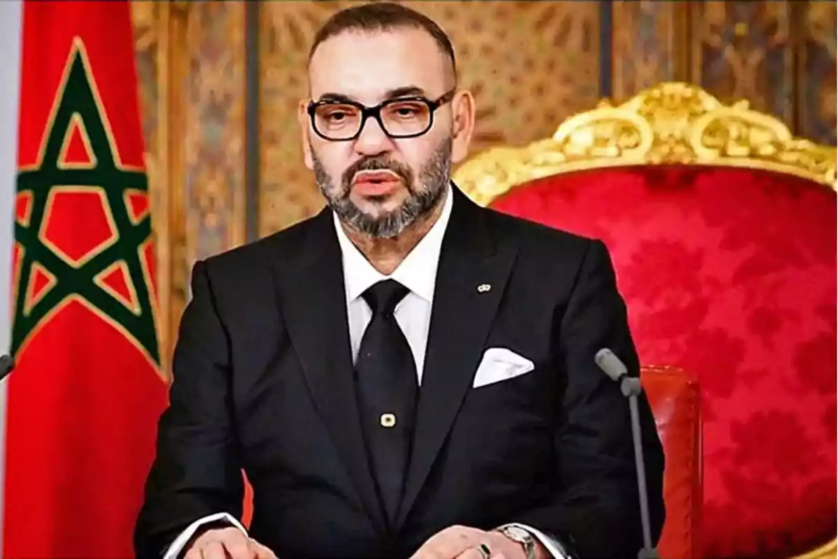 Un hombre con gafas y barba, vestido con un traje negro y corbata, sentado en un trono rojo con un micrófono frente a él y una bandera de Marruecos en el fondo.