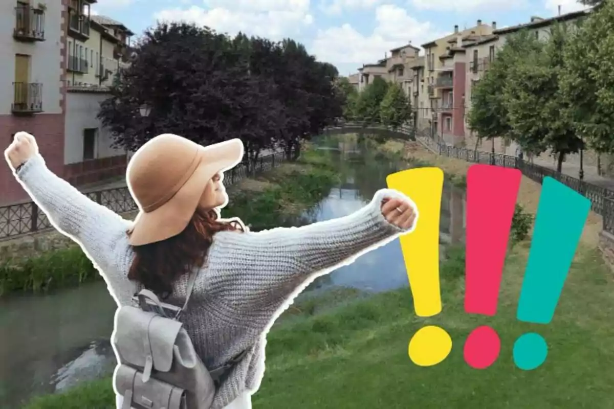 Una mujer con sombrero y mochila levanta los brazos en señal de alegría frente a un río rodeado de edificios y árboles, con tres signos de exclamación coloridos en primer plano.