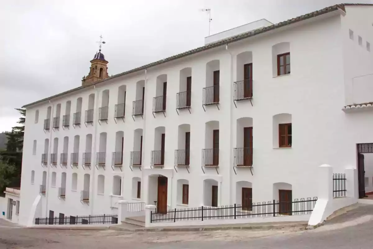 Edificio blanco de tres pisos con múltiples ventanas y balcones en una calle inclinada.