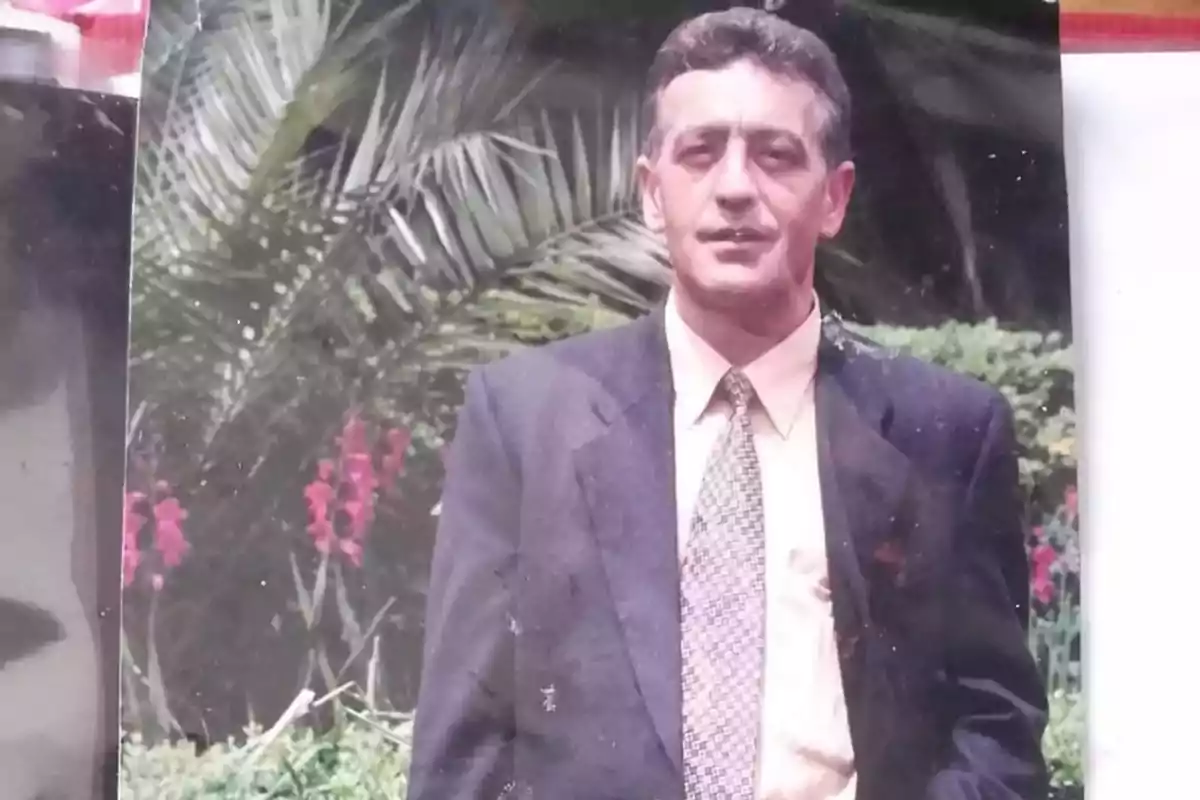 Un hombre con traje y corbata posando frente a un jardín con palmeras y flores.