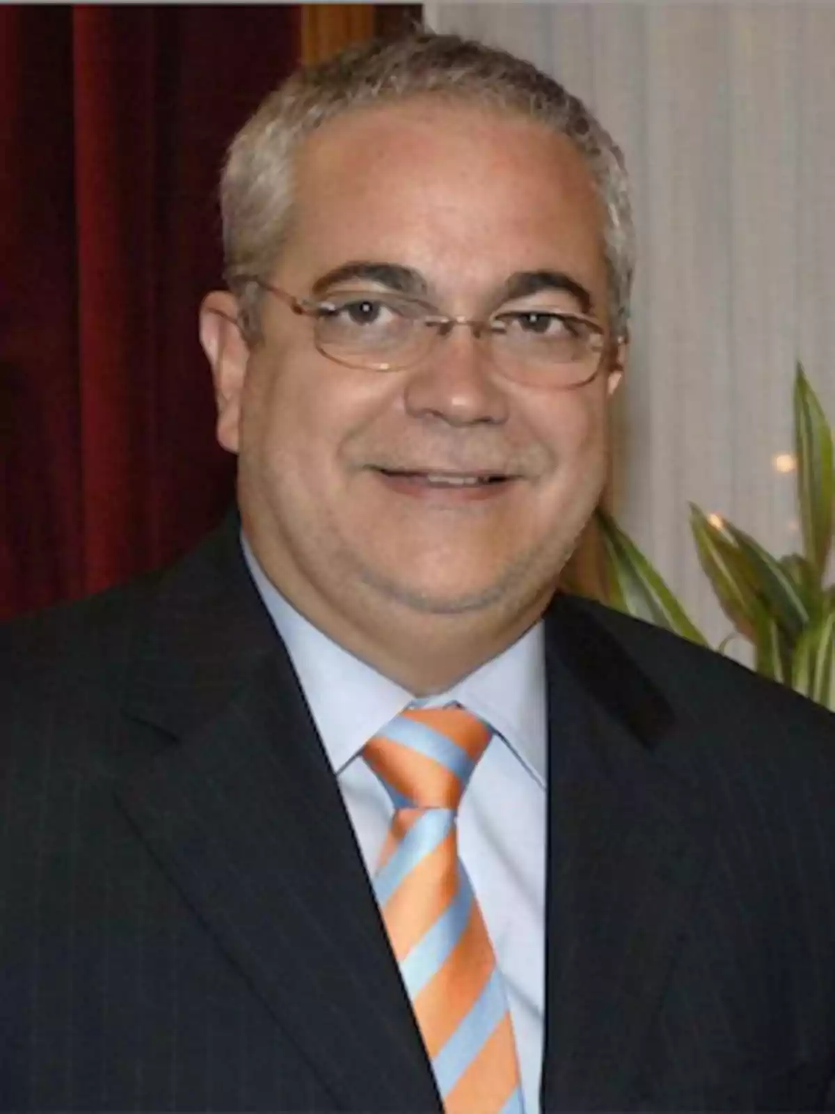 Hombre con gafas y cabello canoso, vestido con traje oscuro y corbata a rayas naranjas y azules, sonriendo frente a una cortina roja y una planta.