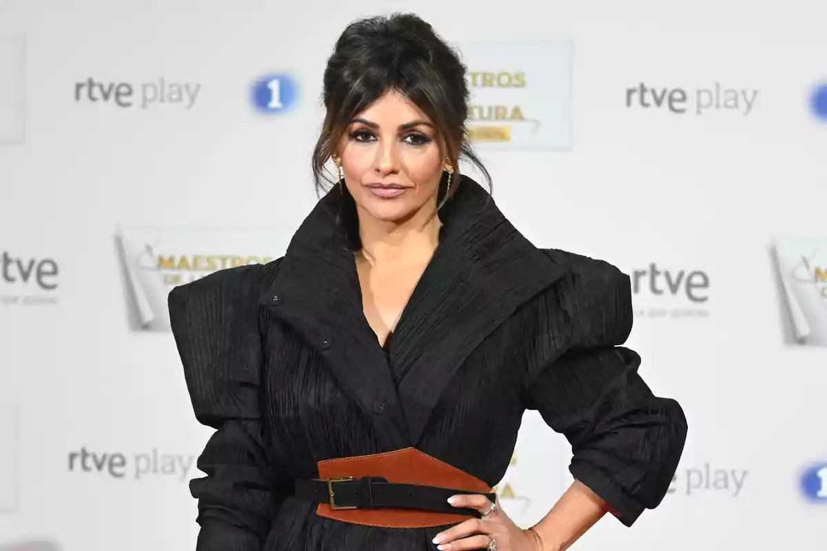 Una persona con un vestido negro elegante y cinturón marrón posando en un evento con un fondo de logotipos de RTVE.