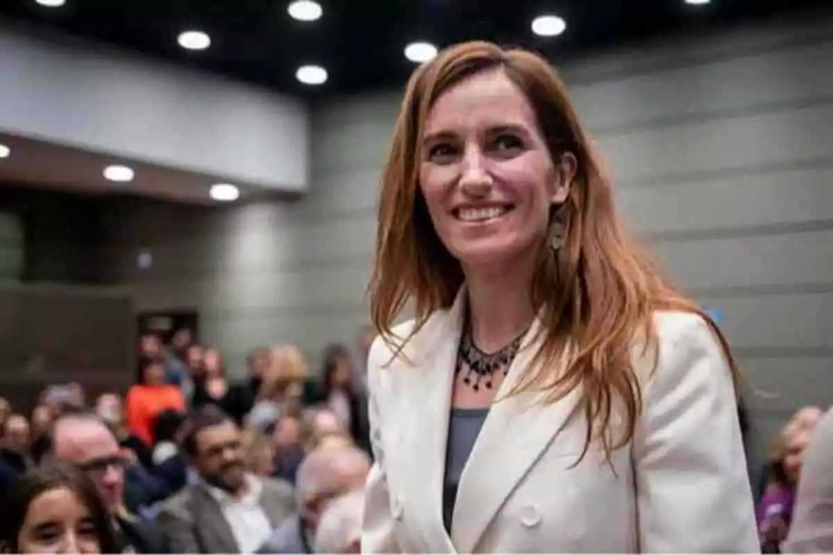 Una mujer con cabello castaño claro y chaqueta blanca sonríe en un evento con público al fondo.