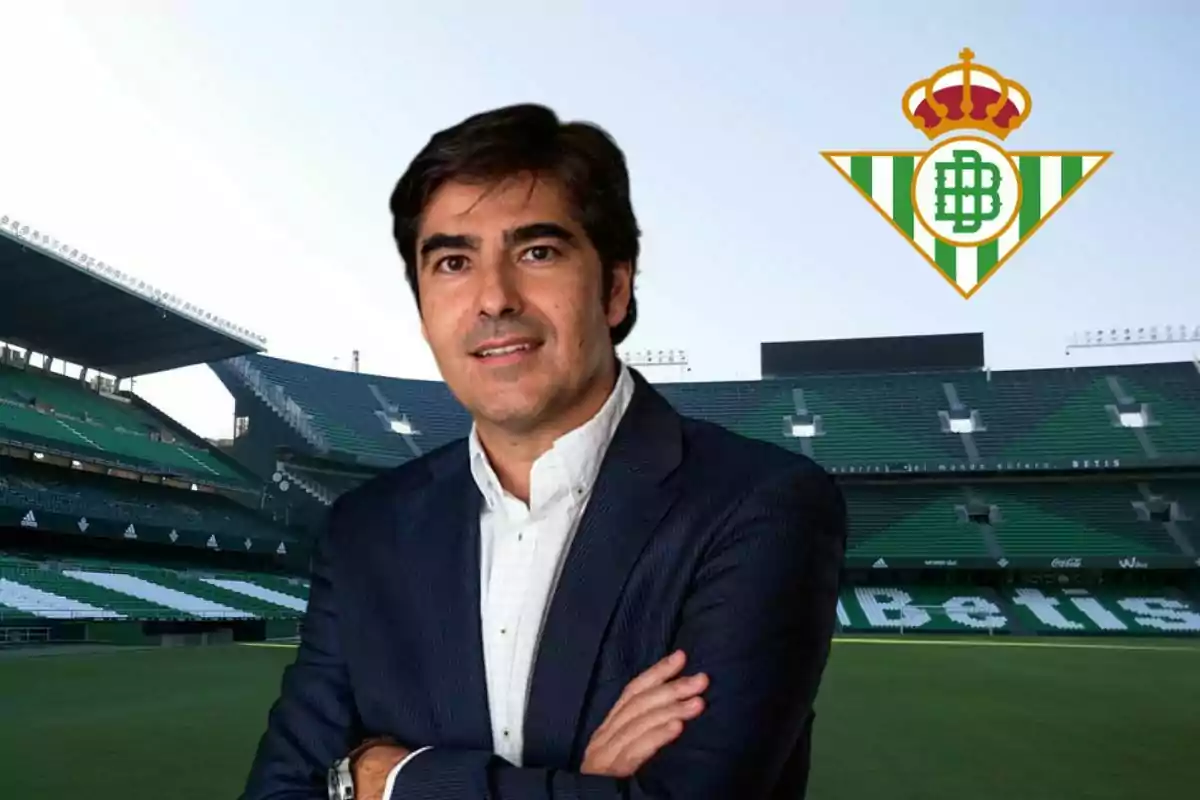 Un hombre con traje posando en un estadio de fútbol con el logo del Real Betis en el fondo.